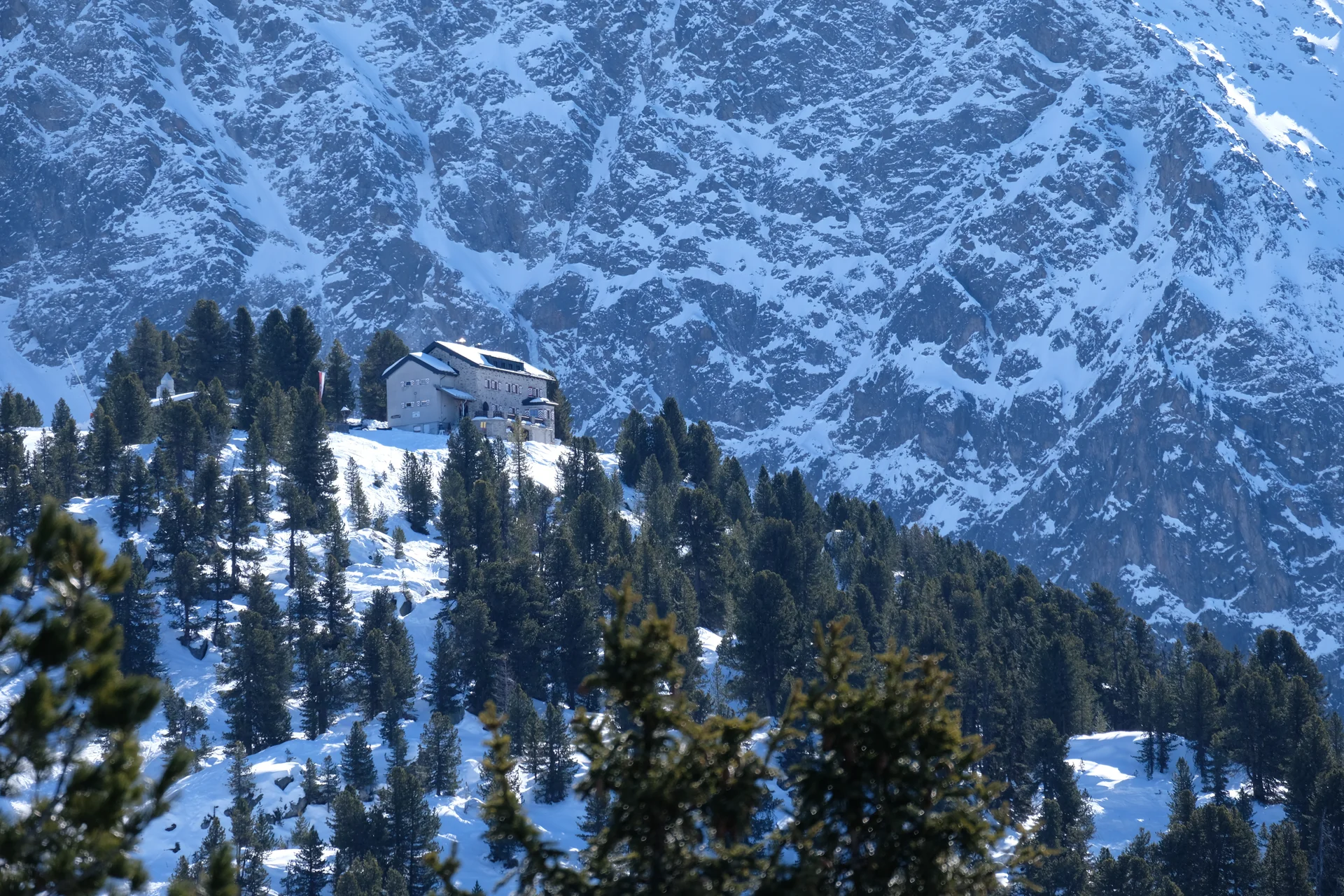 Bielefelder Hütte im Winter. Die Hütte liegt auf einem Hügel, der mit Schnee bedeckt und von der Sonne beschienen ist. Rings herum steht Tannen und Fichten, die grün sind, also keine Schneeauflagen haben. Hinter dem Hügel mit der Hütte sieht man die steil emporragende, teils mit Schnee bedeckte Felswand. Die BIelefelder Hütte sieht gegen die Felswand eher klein aus. | © DAV Bielefeld