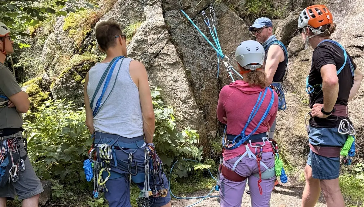 „Teilnehmende eines Kletterkurses besprechen am Einstieg einer Felswand den Aufbau eines Seilsystems. Mit Helmen, Gurten und Sicherungsmaterial ausgestattet bereitet sich die Gruppe auf das Klettern und Abseilen im natürlichen Felsgelände vor.“ | © Gunnar Helms