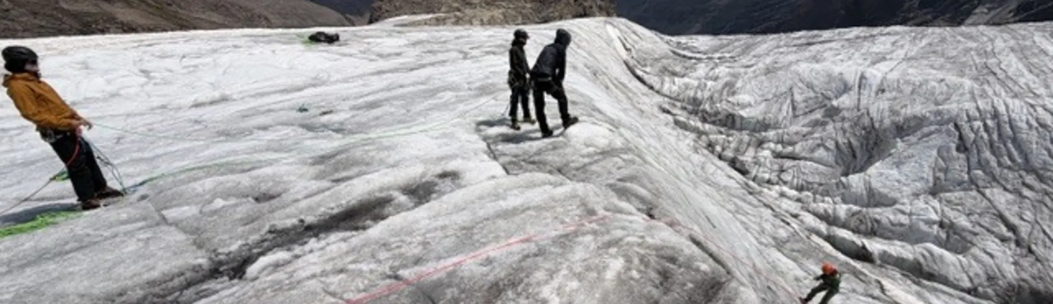 Mehrere Personen bewegen sich angeseilt auf einem Gletscher. Einige befinden sich auf einer steilen Eisfläche, während andere auf einem flacheren Abschnitt stehen. Im Hintergrund sind Berggipfel und Wolken zu sehen. | © P.J.Müller