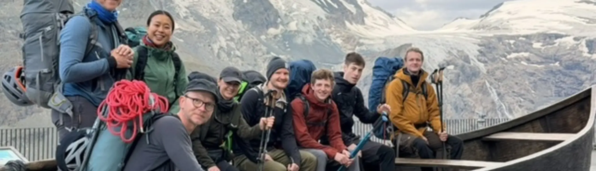 Gruppe von Bergsteigenden mit Rucksäcken und alpiner Ausrüstung sitzt und steht in einem hölzernen Aussichtselement vor einer Gletscherlandschaft und hohen Berggipfeln. | © P.J.Müller