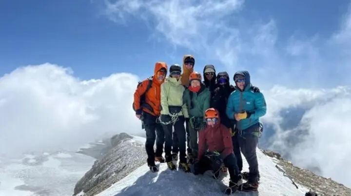 Gruppe von Bergsteigenden mit Helmen, Schutzbrillen und Hochtourenausrüstung auf einem schneebedeckten Gipfelgrat. Im Hintergrund sind Wolken, Gletscherflächen und hochalpines Gelände zu sehen. | © P.J.Müller