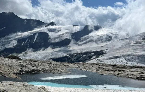Bergsee vor einer Gletscherlandschaft mit schneebedeckten Gipfeln und Wolken. Felsiger Untergrund umgibt das Wasser in hochalpiner Umgebung. | © P.J.Müller