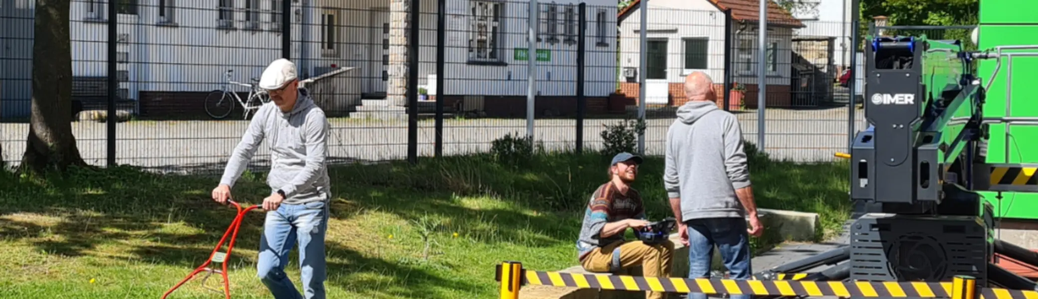 Auf dem Foto sind drei Personen im Freien zu sehen. Links mäht eine Person mit einem kleinen Handrasenmäher das Gras entlang einer niedrigen Steinmauer. Rechts stehen zwei Personen neben einer großen grünen Maschine und unterhalten sich. Der Bereich ist mit gelb-schwarzen Absperrbändern gesichert. Im Hintergrund sieht man ein weißes Gebäude mit rotem Dach, mehrere Fahnen – darunter eine Regenbogenflagge – und Bäume mit frischem Grün. Der Himmel ist klar und blau, es scheint ein sonniger Tag zu sein. | © Thomas Fromme