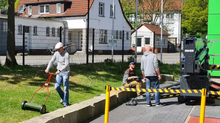 Auf dem Foto sind drei Personen im Freien zu sehen. Links mäht eine Person mit einem kleinen Handrasenmäher das Gras entlang einer niedrigen Steinmauer. Rechts stehen zwei Personen neben einer großen grünen Maschine und unterhalten sich. Der Bereich ist mit gelb-schwarzen Absperrbändern gesichert. Im Hintergrund sieht man ein weißes Gebäude mit rotem Dach, mehrere Fahnen – darunter eine Regenbogenflagge – und Bäume mit frischem Grün. Der Himmel ist klar und blau, es scheint ein sonniger Tag zu sein. | © Thomas Fromme
