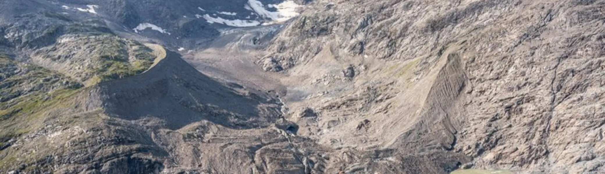 Eine Gruppe von Wandernden geht auf einem schmalen Bergpfad in einer alpinen Landschaft. Im Hintergrund ragen hohe, felsige Berge mit Schneefeldern und einem Gletscher auf. Unten im Tal liegt ein kleiner Gletschersee mit hellgrünem Wasser. Die Personen tragen Rucksäcke und Wanderstöcke und bewegen sich bergab bei sonnigem Wetter. | © DAV / Julian Rohn