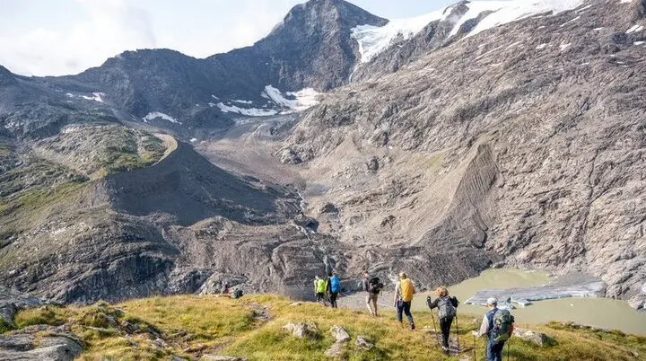 Eine Gruppe von Wandernden geht auf einem schmalen Bergpfad in einer alpinen Landschaft. Im Hintergrund ragen hohe, felsige Berge mit Schneefeldern und einem Gletscher auf. Unten im Tal liegt ein kleiner Gletschersee mit hellgrünem Wasser. Die Personen tragen Rucksäcke und Wanderstöcke und bewegen sich bergab bei sonnigem Wetter. | © DAV / Julian Rohn