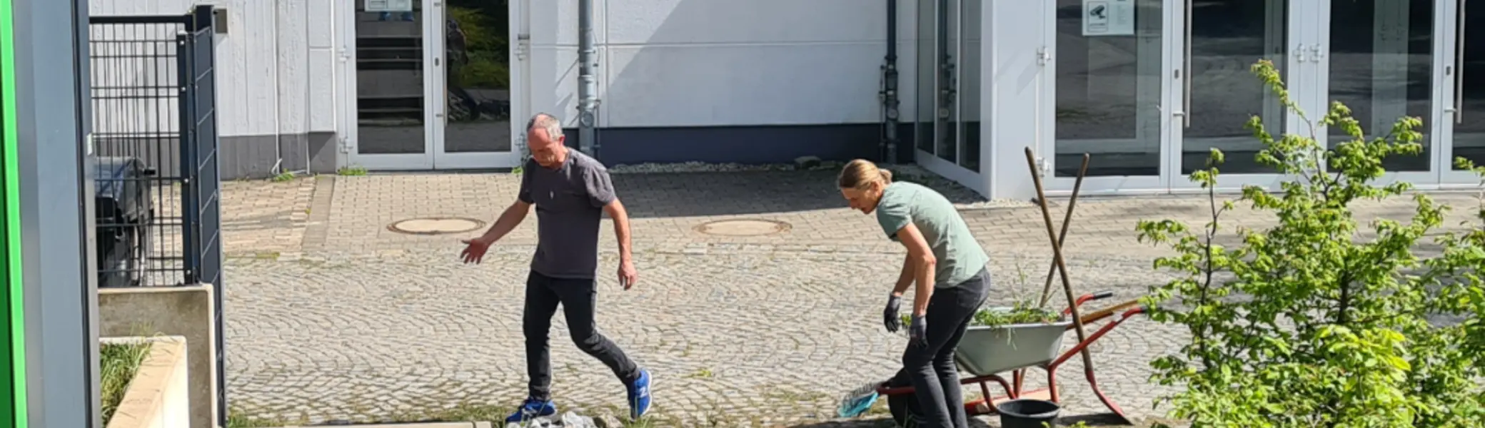    Zwei Personen arbeiten im Freien vor einem weißen Gebäude mit der Aufschrift „Regine-Hildebrandt-Halle“ und „DAV Bielefeld“. Eine Person steht auf einem gepflasterten Weg, die andere arbeitet mit einer Schubkarre und Werkzeugen neben grünen Sträuchern. Im Hintergrund sind Glas­türen und ein Schild mit „Stiftung Kultur Kommunikation Sieker“ zu sehen. | © Thomas Fromme