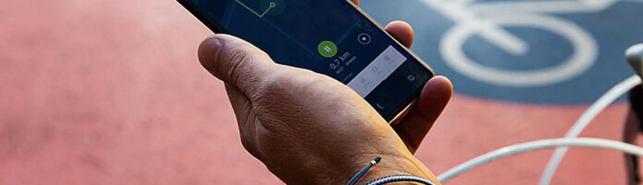 Eine Person hält ein Smartphone in der Hand, auf dessen Bildschirm eine Navigations-App mit einer grünen Route angezeigt wird. Im Hintergrund ist ein roter Fahrradweg mit einem weißen Fahrradsymbol zu sehen, und der Lenker eines Fahrrads ragt ins Bild. | © Klima Bündnis Services / Felix Krammer