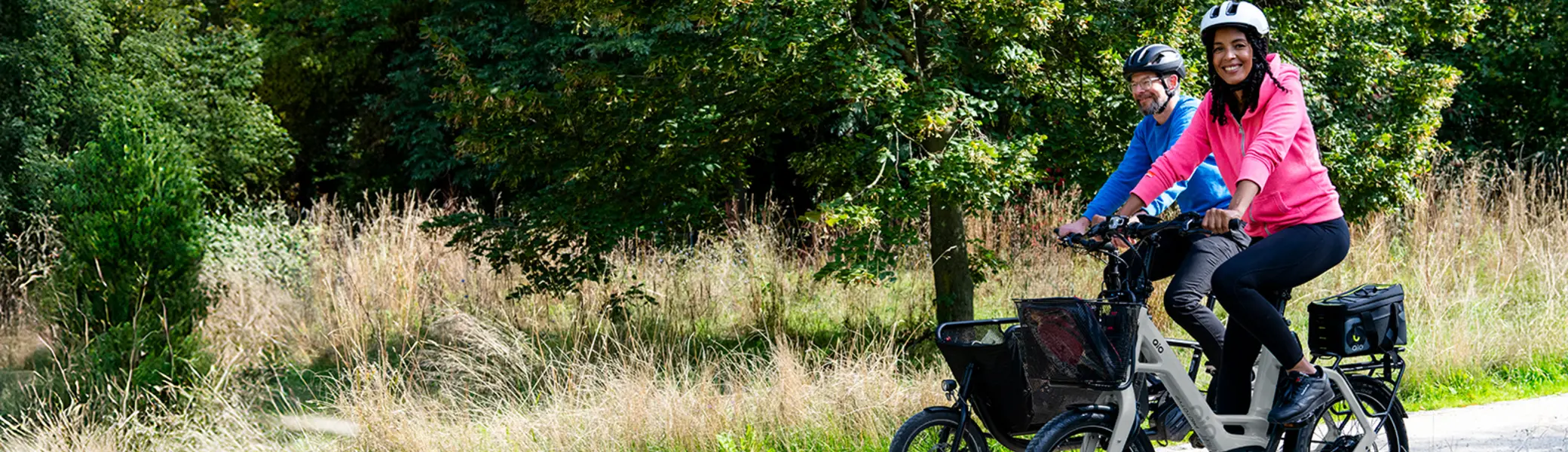 Zwei Personen fahren auf einem hellen, geschotterten Weg durch eine grüne Landschaft mit hohen Bäumen und Gras. Beide tragen Helme und Freizeitkleidung. Die vordere Person trägt einen pinken Pullover und lächelt, die hintere Person trägt einen blauen Pullover. Beide fahren auf modernen E-Bikes mit Gepäcktaschen. | © Klima Bündnis Services / Felix Krammer