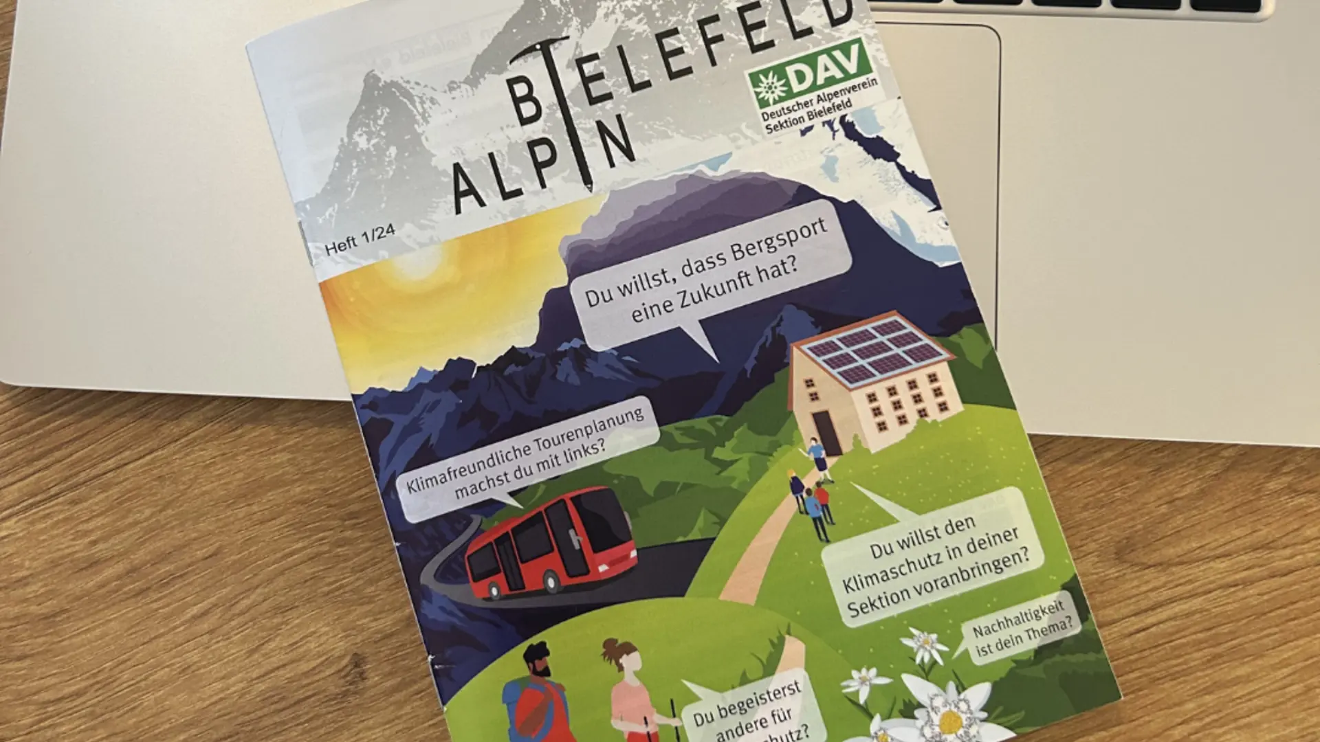 Ein Magazin mit dem Titel „Bielefeld Alpin“ liegt auf einer Laptop-Tastatur. Auf dem Cover ist eine farbige Illustration mit Bergen, einer Seilbahn, einem Haus mit Solaranlage und Wandernden zu sehen. Mehrere Sprechblasen enthalten Texte zu Klimaschutz und nachhaltigem Bergsport. Oben rechts steht das Logo „DAV – Deutscher Alpenverein Sektion Bielefeld“. | © Nicole Laska
