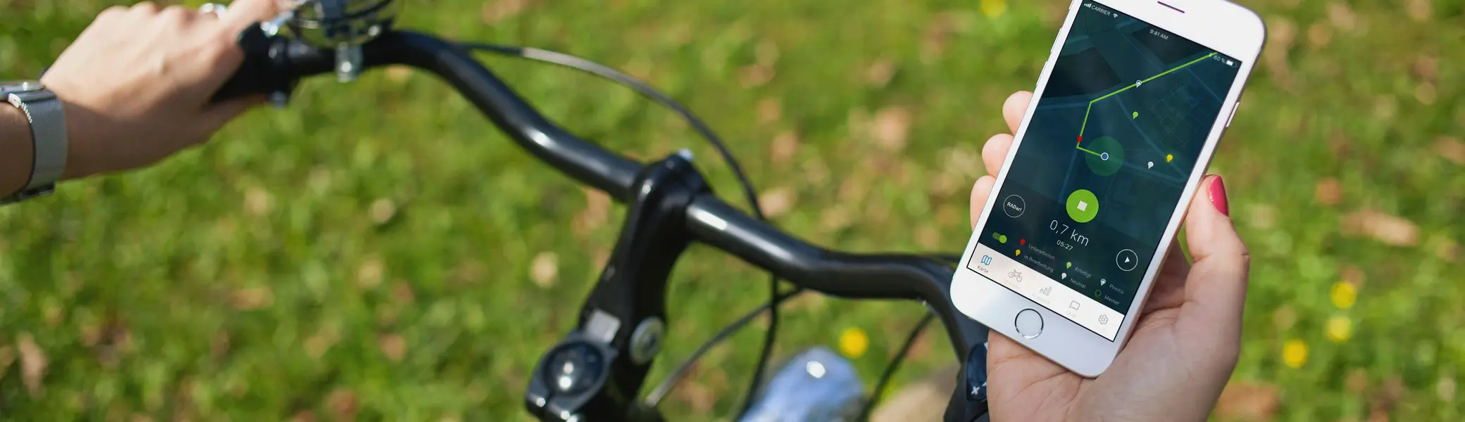 Eine Person hält ein Smartphone in der rechten Hand, während sie Fahrrad fährt. Auf dem Bildschirm ist eine Navigations-App mit einer grünen Route und der Anzeige „0,7 km“ zu sehen. Die linke Hand greift den Fahrradlenker mit einer silbernen Klingel. Im Hintergrund ist eine grüne Wiese mit kleinen gelben Blumen. | © Klima Bündnis Services / Felix Krammer