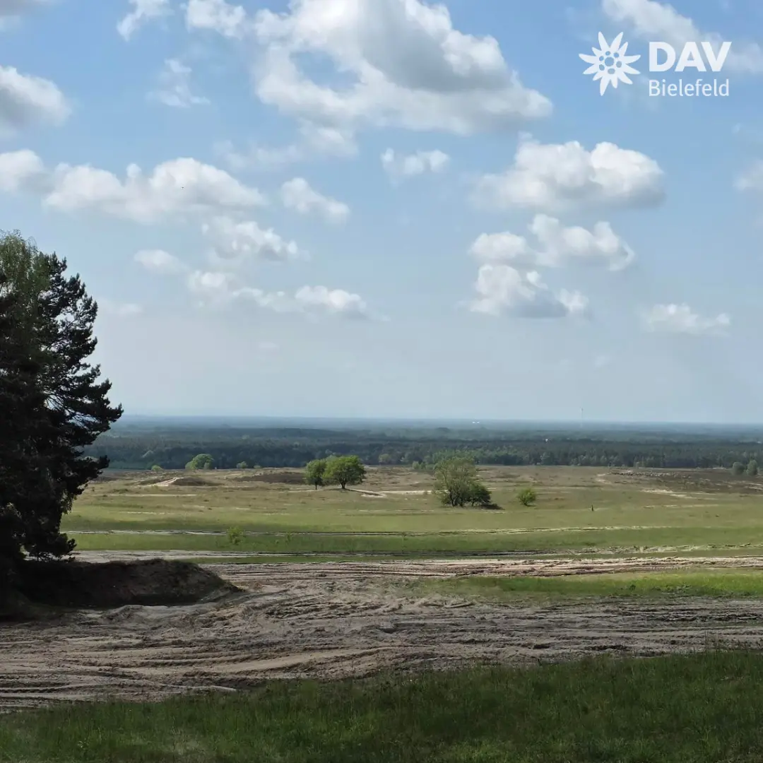 Weite Landschaft mit sandigen Flächen im Vordergrund und grünen Wiesen dahinter. Einzelne Bäume stehen verstreut, am Horizont ist ein dichter Wald zu sehen. Der Himmel ist hellblau mit weißen, lockeren Wolken. Links im Bild ragt eine dunkle Baumkrone ins Bild. Oben rechts befindet sich das Logo „DAV Bielefeld“. | © Bianca Brühöfener
