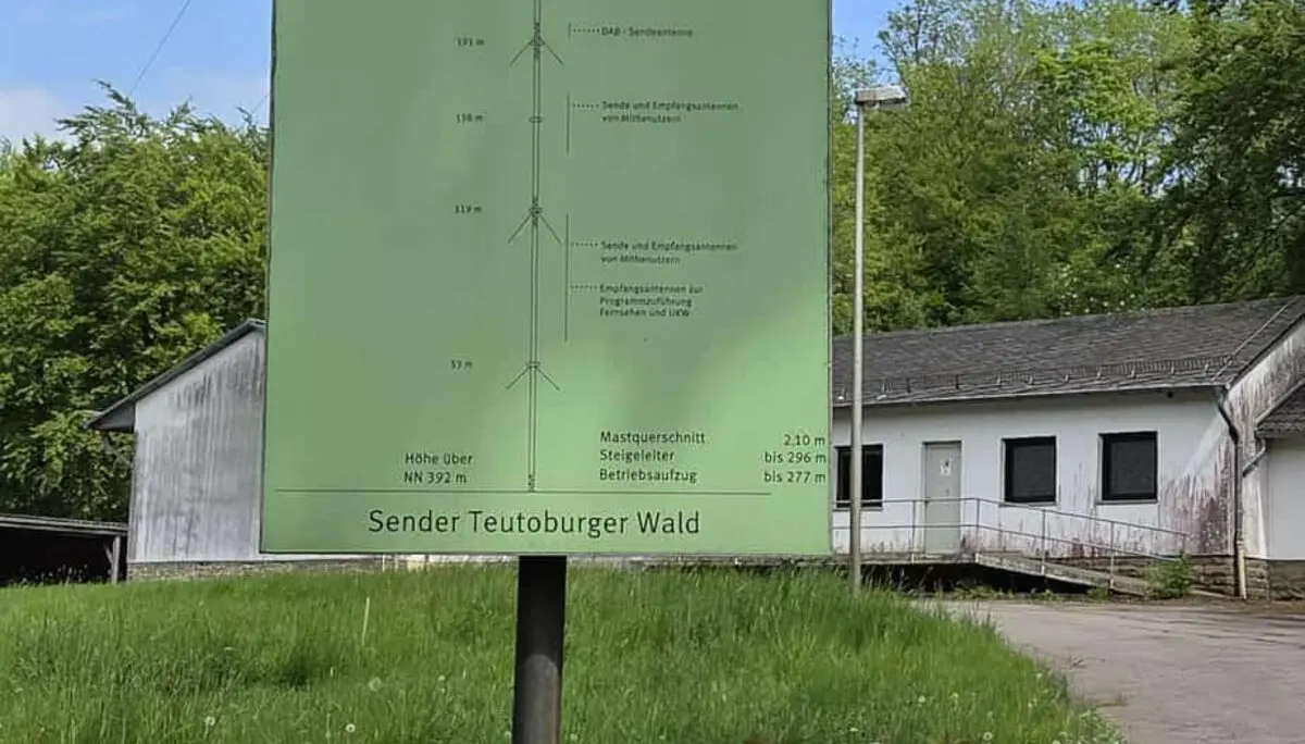 Informationsschild des WDR zum Sender Teutoburger Wald auf einer Wiese neben Gebäuden und Bäumen unter blauem Himmel. | © Bianca Brühöfner