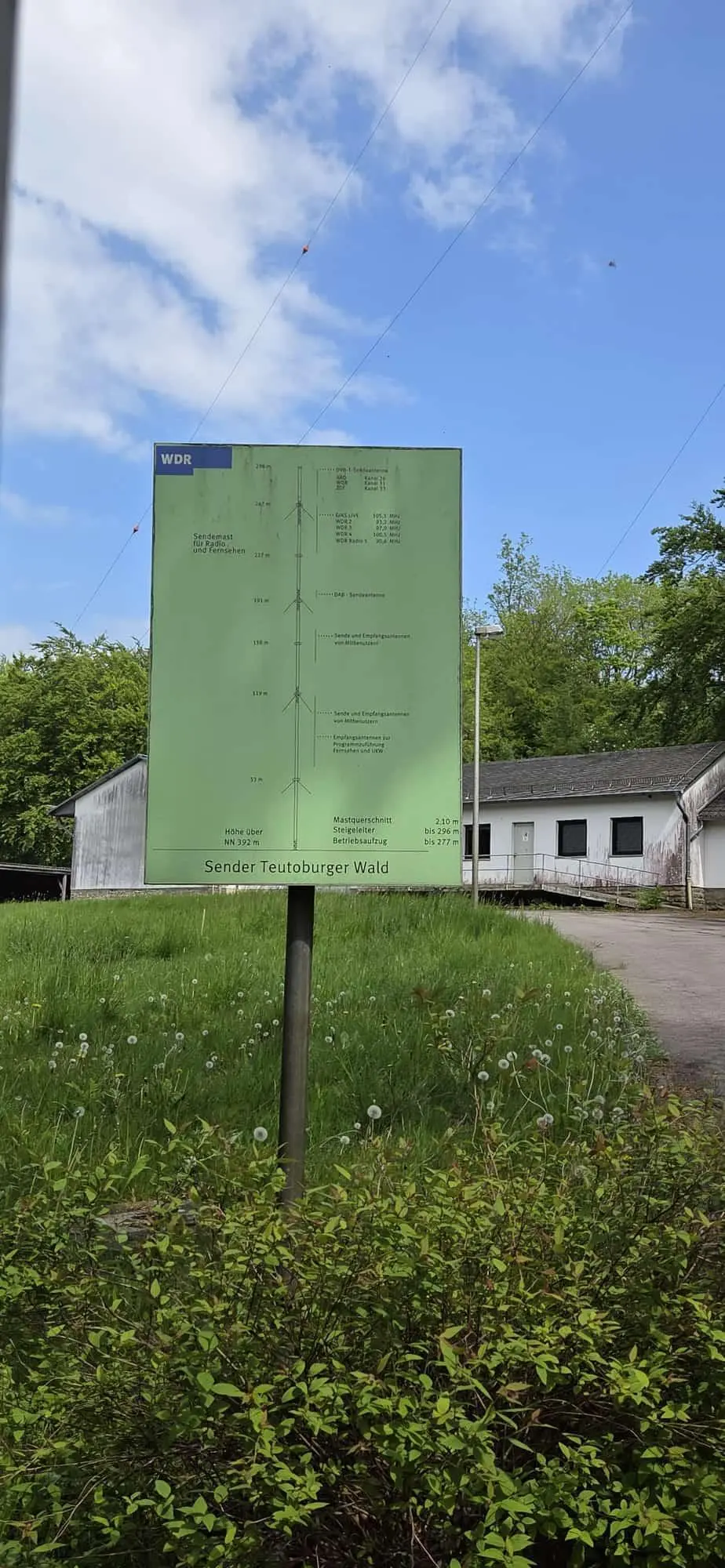 Informationsschild des WDR zum Sender Teutoburger Wald auf einer Wiese neben Gebäuden und Bäumen unter blauem Himmel. | © Bianca Brühöfner