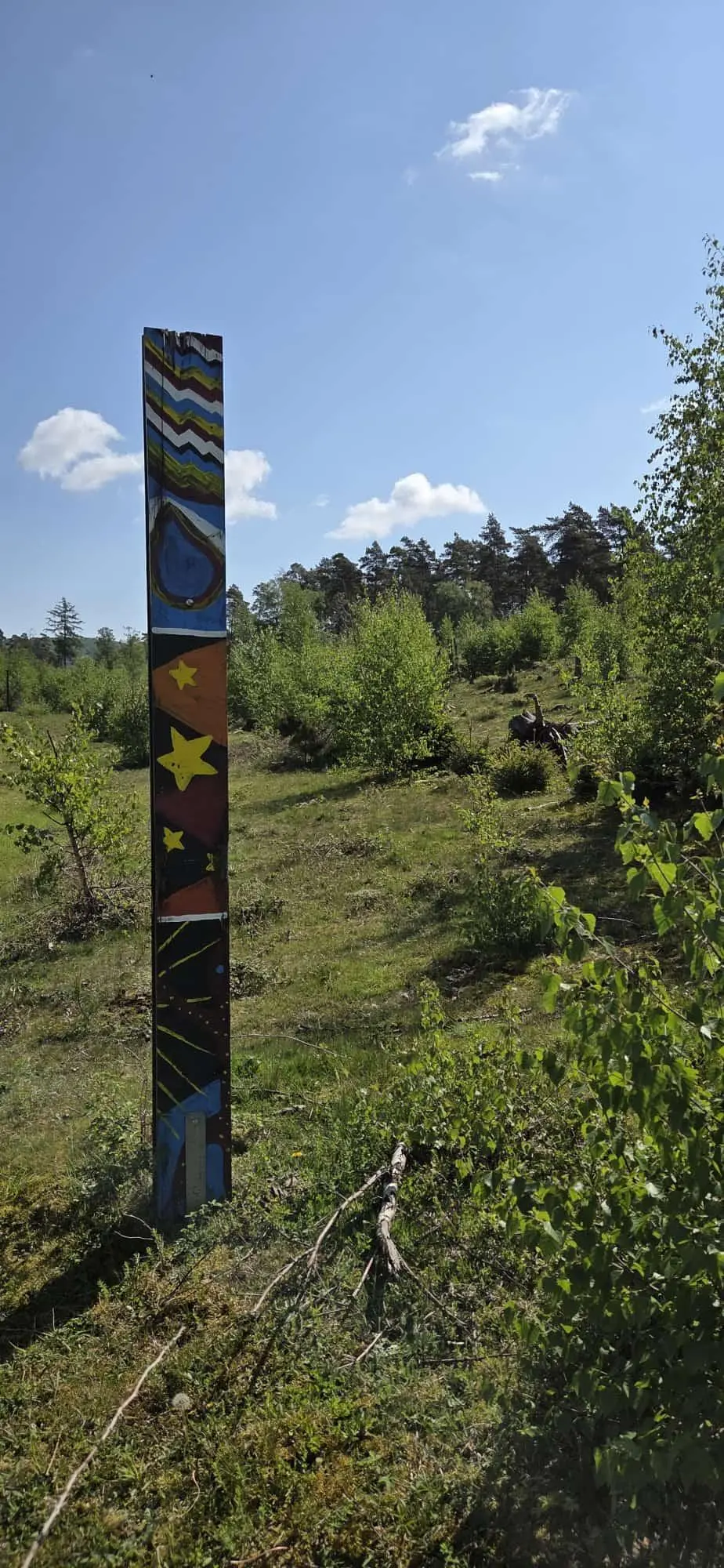 Bunte, bemalte Holzstele steht auf einer Wiese mit Sträuchern vor einem Wald unter blauem Himmel. | © Bianca Brühöfner