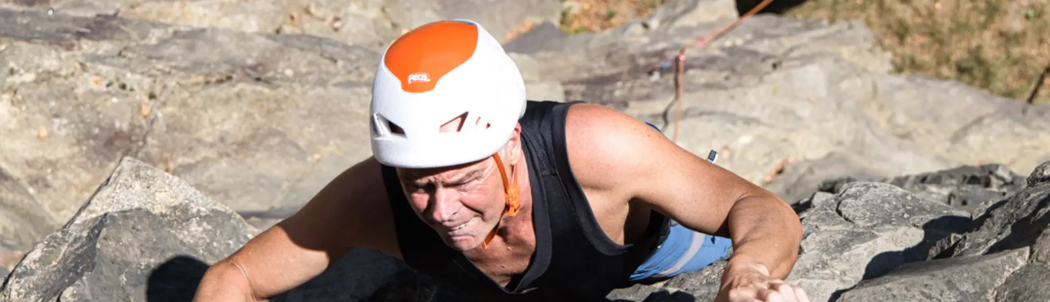 Person mit Kletterhelm klettert an einer Felswand nach oben. Im Hintergrund sind am Wandfuß ausgelegte Kletterseile und weitere Personen zu sehen. | © Thorsten Neuenbäumer