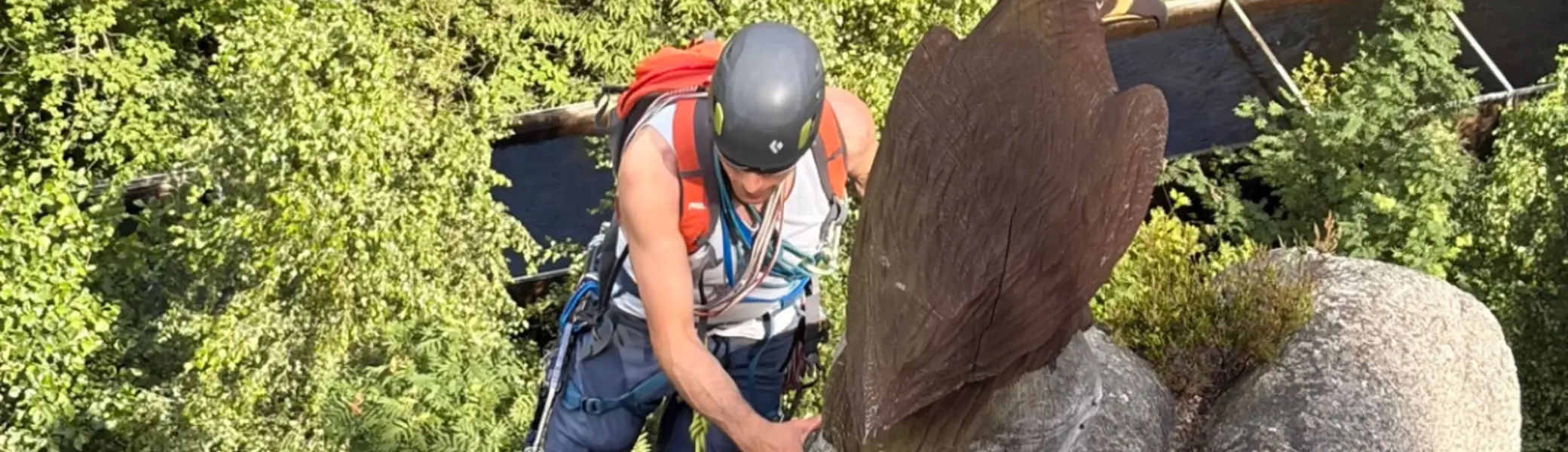 Kletternde Person mit Helm und Rucksack erreicht den Ausstieg einer Felsformation. Neben der Route steht eine hölzerne Adlerfigur, im Hintergrund ist ein bewaldetes Tal zu sehen. | © Gunnar Helms